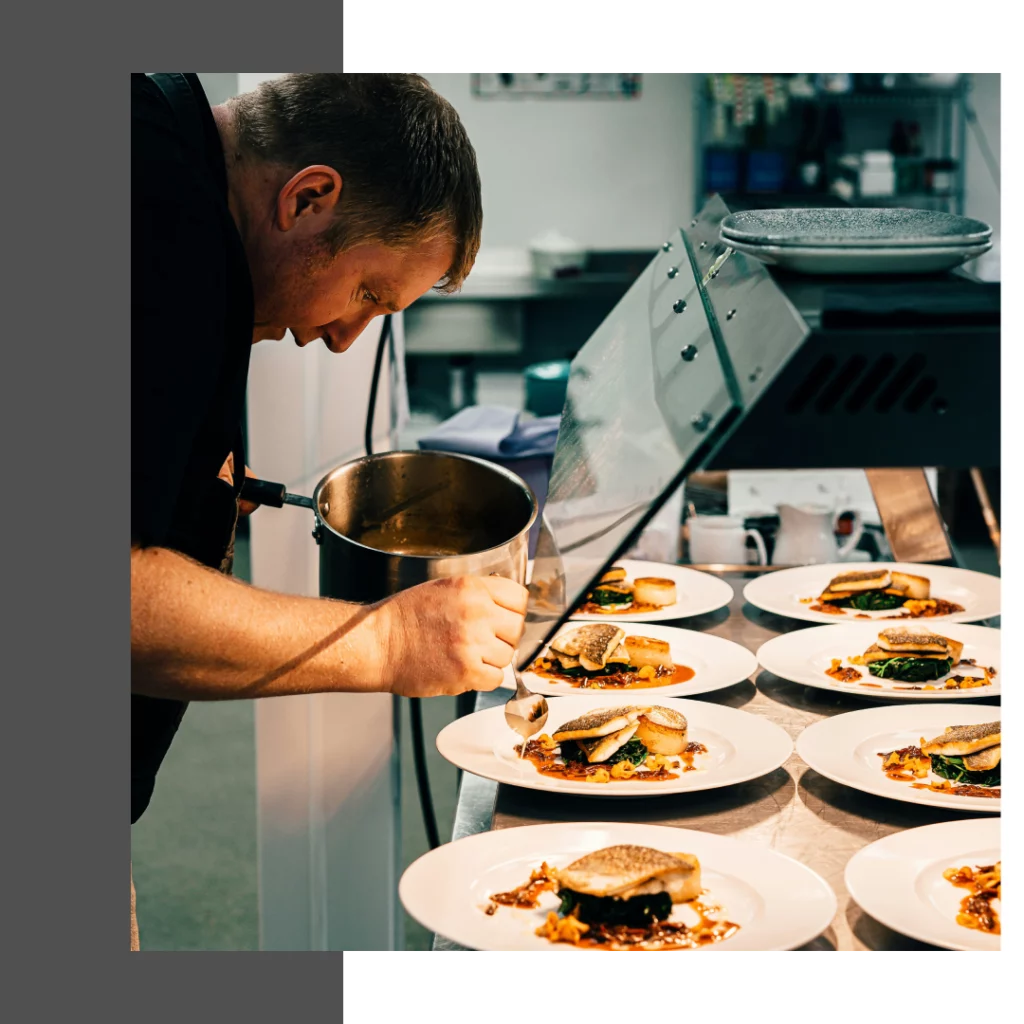A male chef, wearing a black shirt, is focused on garnishing a row of plated dishes. He holds a silver saucepan in his left hand and a spoon in his right, carefully drizzling sauce onto each plate. The dishes are identical, featuring a piece of fish on a bed of greens.